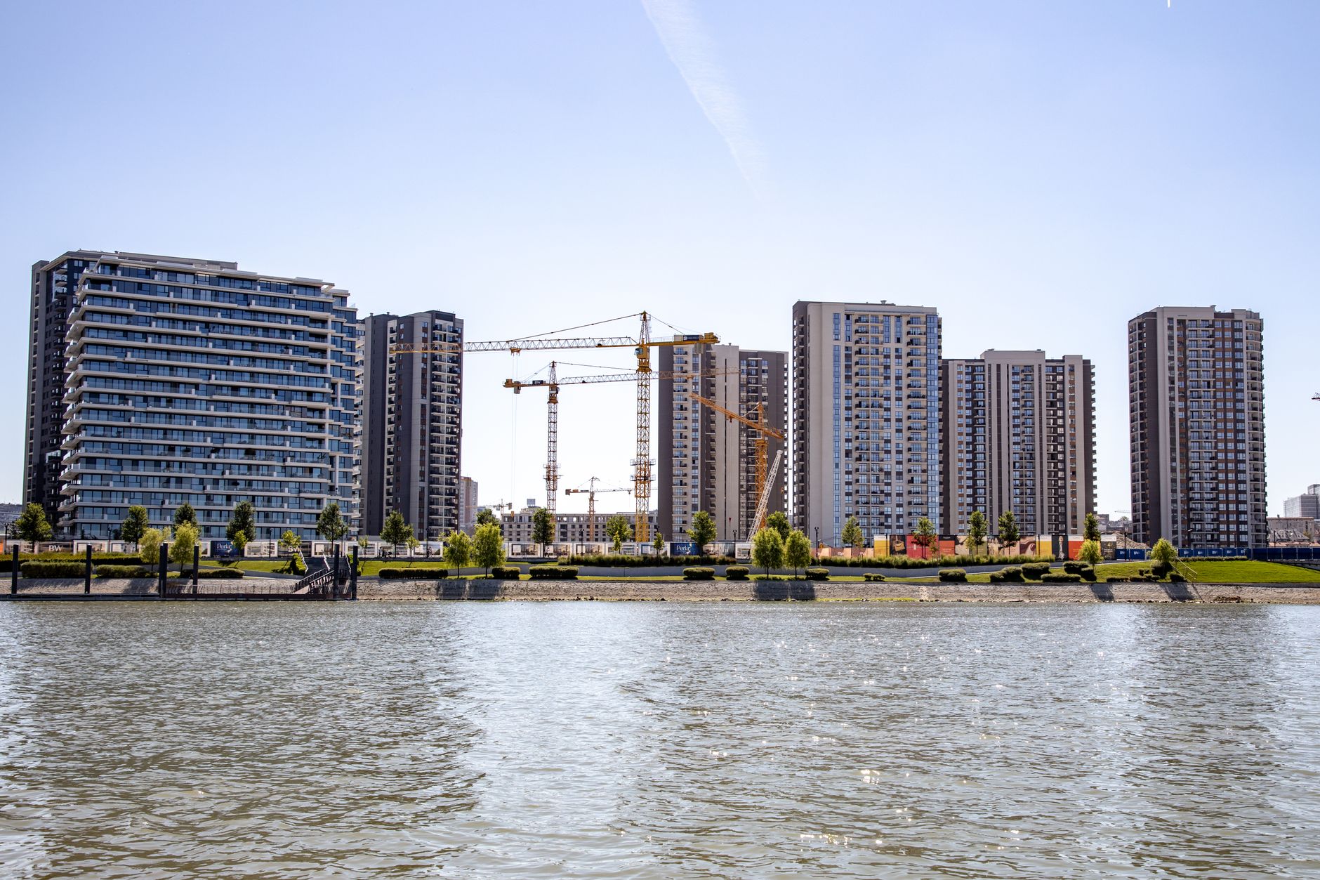 Aerial view of a high-rise luxury residential tower under construction along a city waterfront, cranes visible against a clear sky