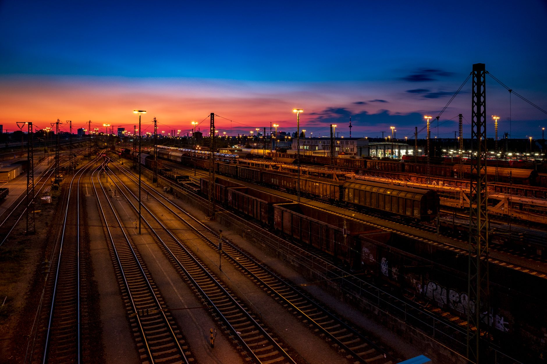 exterior of a modern rail yard at dusk, with lines of freight cars extending toward the horizon under an orange sky and signal lights glowing along the tracks